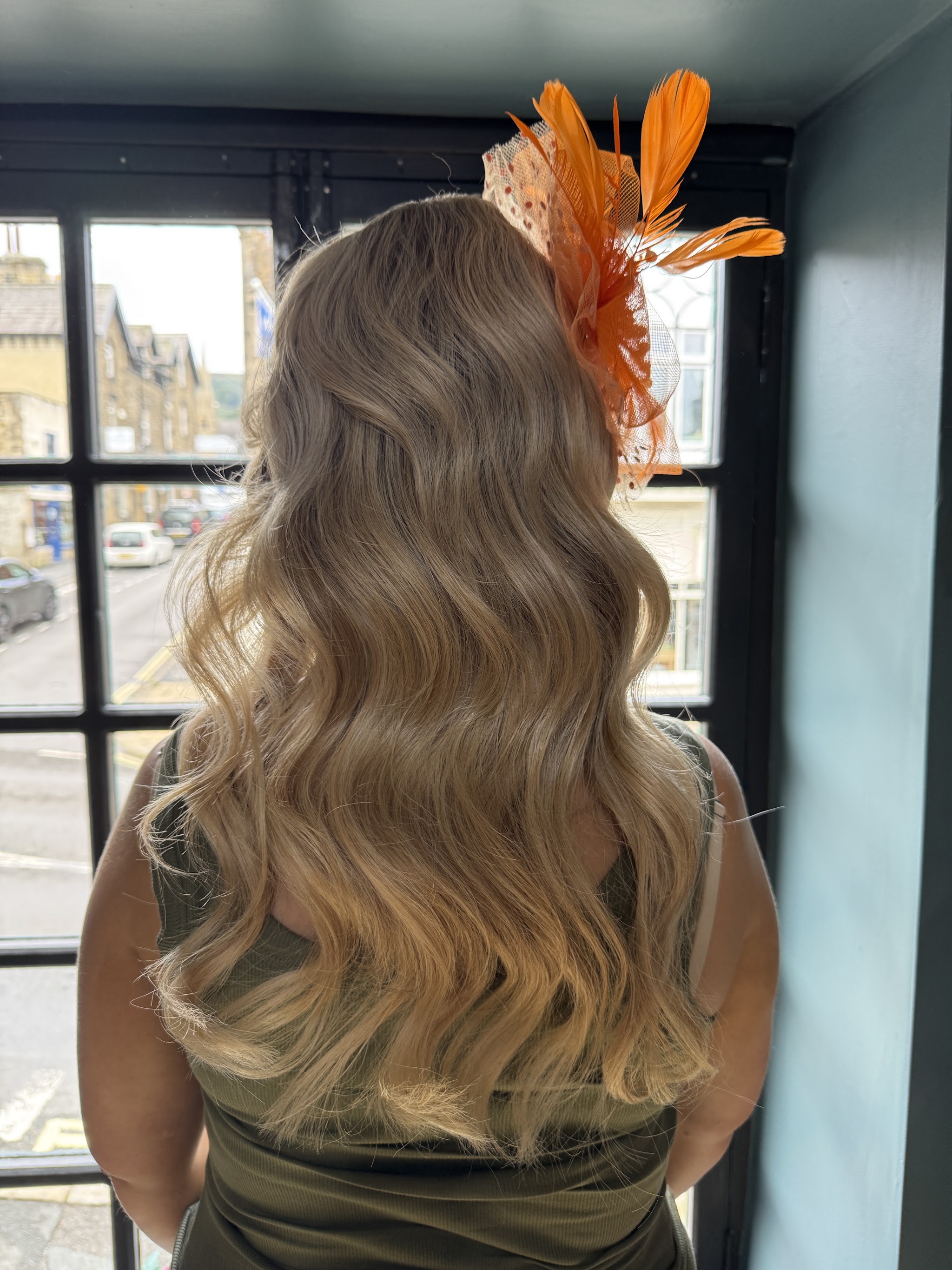 Long blonde waves finished with an orange fascinator, photographed by a salon window